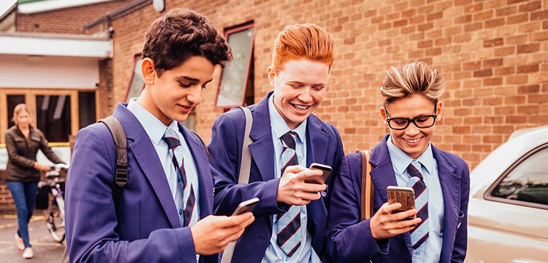 Three high school students walk beside each other looking at their phones and laughing.